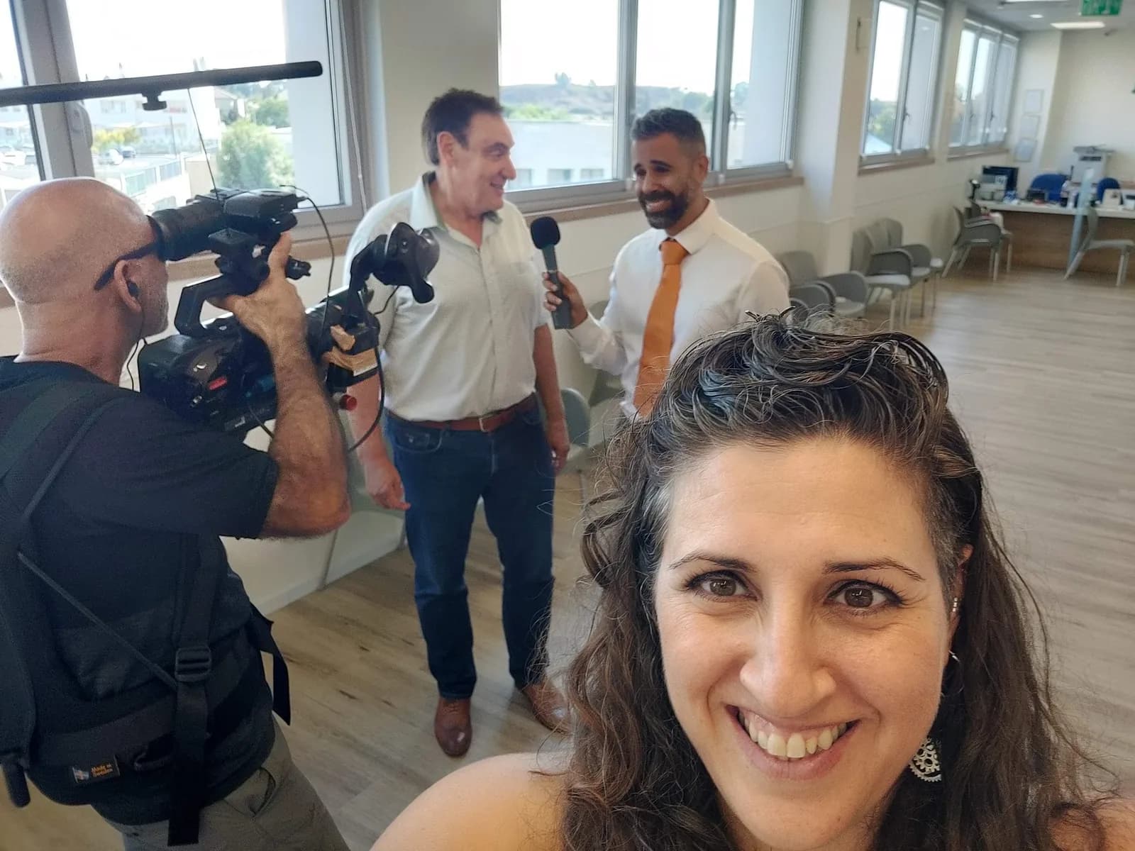 ריאיון עם מיקרופון כף יד באולם מואר, צלם עם מצלמת כתף ומיי מיי סגל בחזית